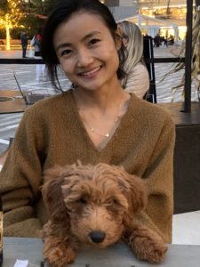 Yeen, wearing a brown sweater and smiling, is seated and holding Milo, a brown fluffy dog.