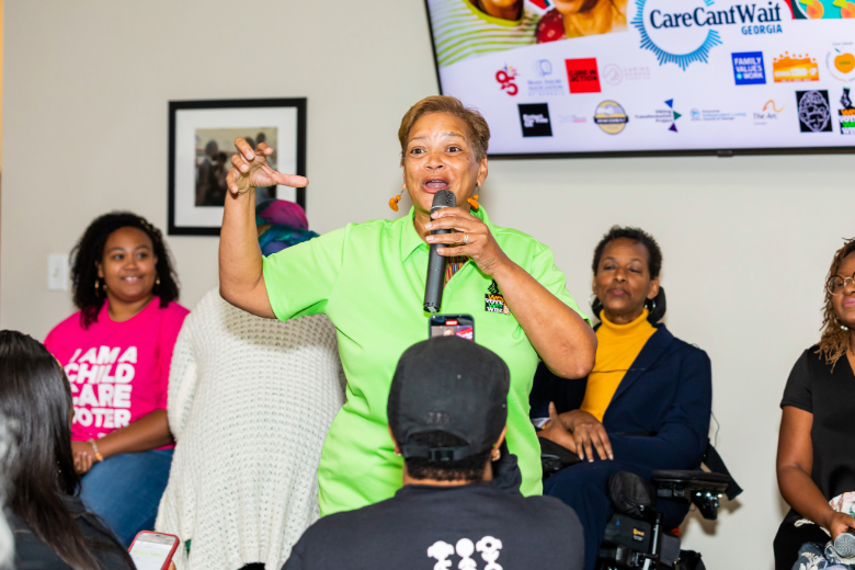 A person with light brown skin stands and speaks animatedly into a microphone at a Georgia roundtable event.