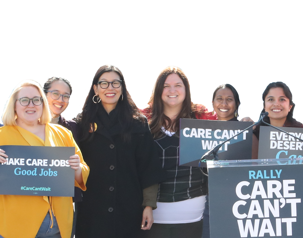 A group of Caring Across staff members, including Ai-jen Poo, smile and hold signs that read