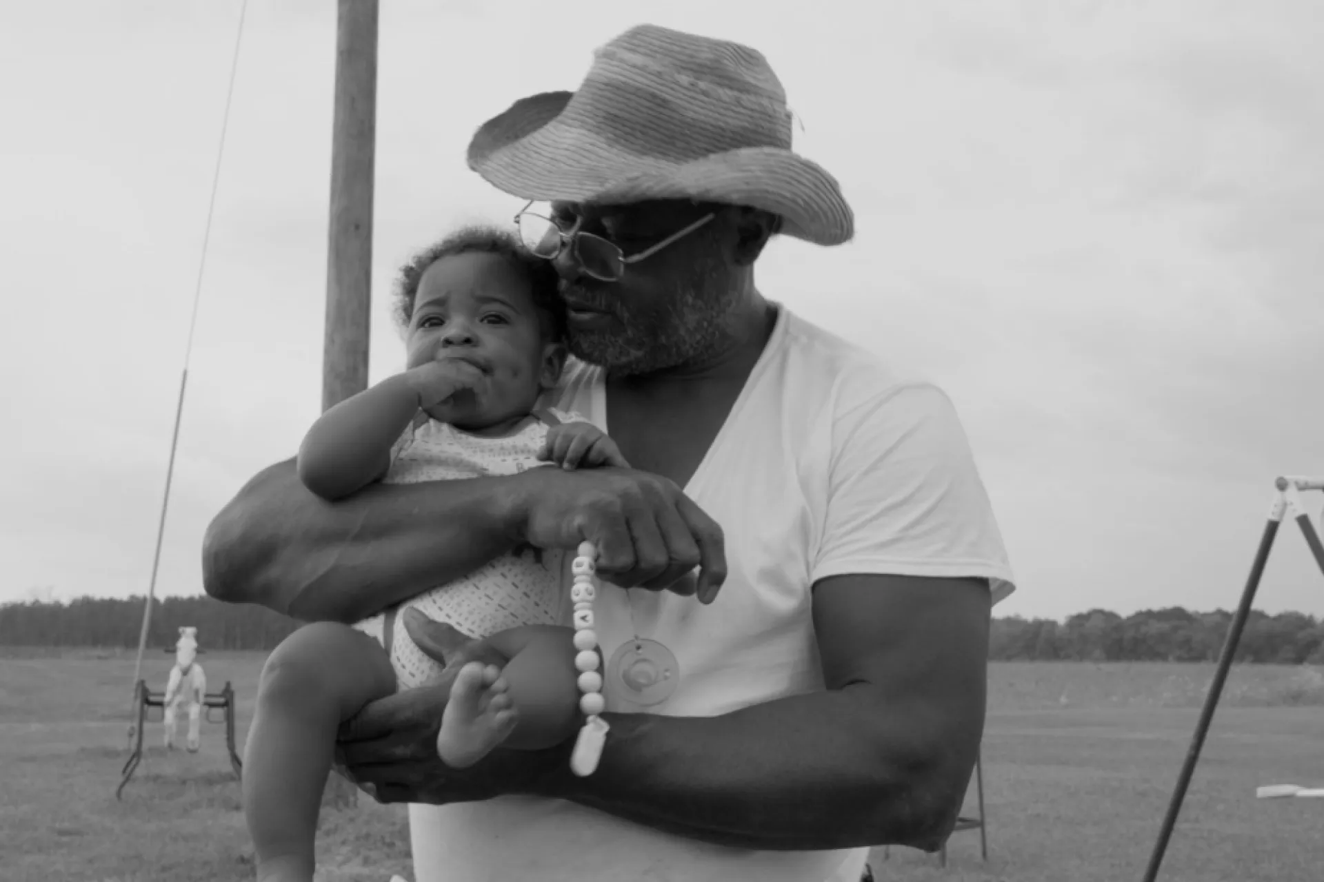 Black and white image of an older Black man holding a Black baby while grabbing a pacifier. The older man is wearing a hat, wire-rimmed glasses and a white t-shirt while the baby is wearing a onesie.